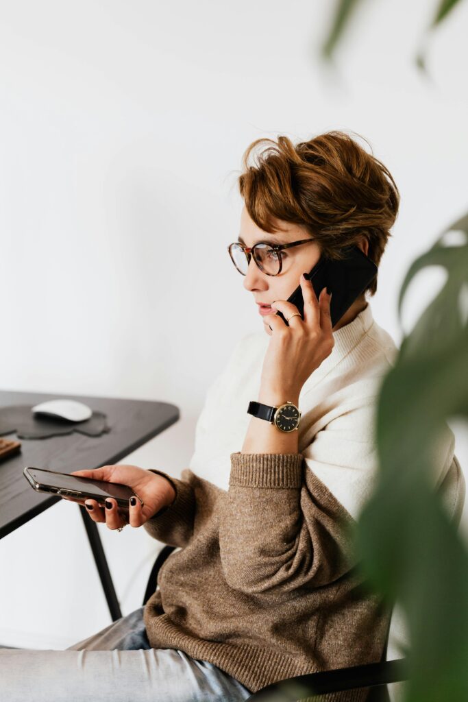 Side view serious female in casual wear and eyeglasses sitting on chair and having conversation via mobile phone in against white wall